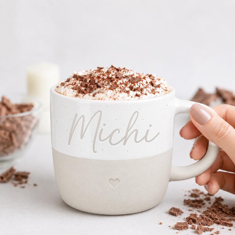 K&ouml;nnte beinhalten: Ein zweifarbiger Keramikbecher mit einer wei&szlig; gesprenkelten oberen H&auml;lfte und einer einfarbigen hellbeigen unteren H&auml;lfte. Das Wort Michi ist in Schreibschrift auf der Vorderseite aufgedruckt, darunter befindet sich ein kleines Herzsymbol. Der Becher ist mit Sahne gef&uuml;llt.