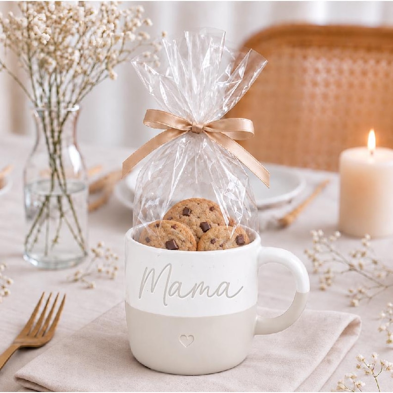 K&ouml;nnte beinhalten: Ein zweifarbiger Keramikbecher mit einem wei&szlig;en oberen Teil, der das Wort Mama in Schreibschrift zeigt, und einem beigen unteren Teil mit einem kleinen Herzsymbol. Der Becher ist mit Schokoladenkeksen in einem transparenten Beutel gef&uuml;llt.