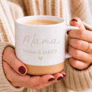 Könnte beinhalten: Ein weißer Keramikbecher mit gesprenkelter Oberfläche, der den geprägten Schriftzug Mama, die Namen NOAH & EMELY sowie ein kleines Herzsymbol zeigt. Der untere Teil des Bechers hat einen glatten, unglasierten beigen Boden und einen Henkel.