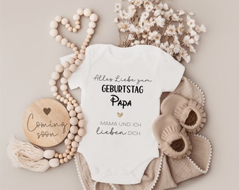 May include: A white baby onesie with the text "Alles Liebe zum Geburtstag Papa Mama und ich lieben dich" in black lettering with a gold glitter heart. The onesie is surrounded by a wooden bead garland, a wooden sign that says "Coming soon" with a heart, a beige blanket, and dried flowers.