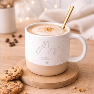Pode incluir: Uma caneca de cer&acirc;mica branca salpicada com uma base bege, cheia de uma bebida e coberta com uma pitada de especiarias. A caneca tem "Mrs. SCHUBER" impressa e est&aacute; sobre um descanso de madeira com biscoitos com pepitas de chocolate.