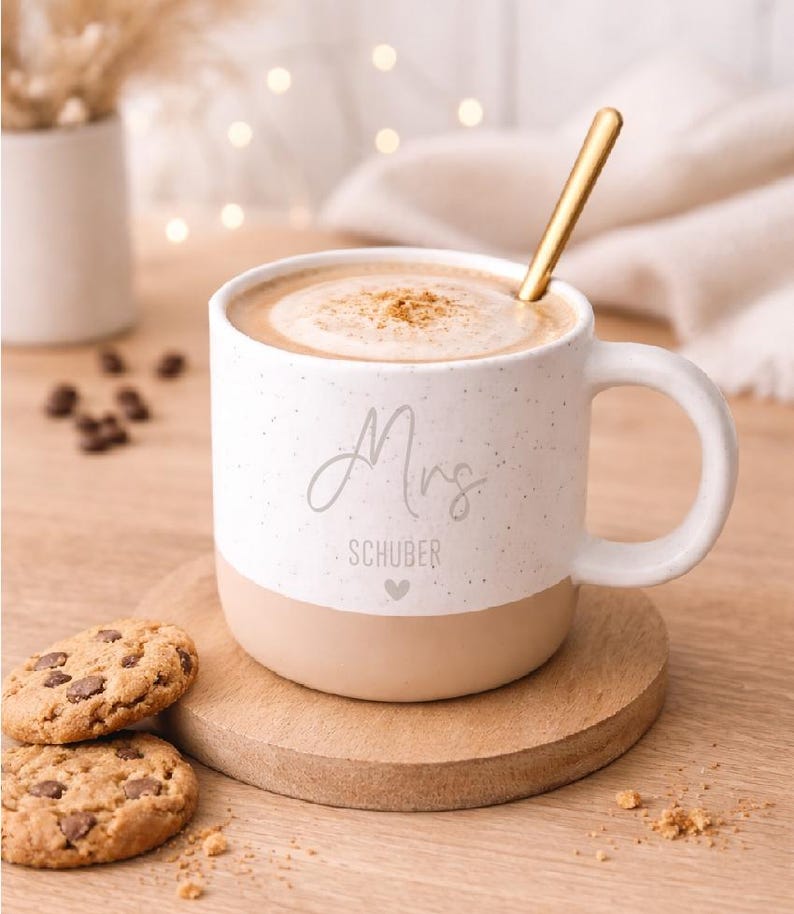 Pode incluir: Uma caneca de cer&acirc;mica branca salpicada com uma base bege, cheia de uma bebida e coberta com uma pitada de especiarias. A caneca tem "Mrs. SCHUBER" impressa e est&aacute; sobre um descanso de madeira com biscoitos com pepitas de chocolate.