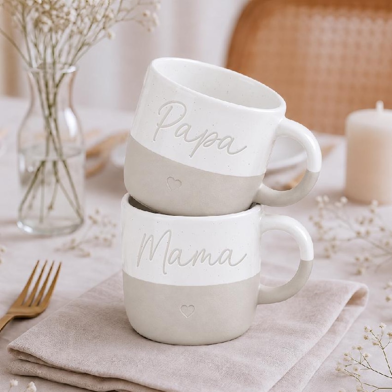 K&ouml;nnte beinhalten: Zwei gestapelte Keramikbecher mit zweifarbigem Design. Die obere H&auml;lfte ist wei&szlig; mit gesprenkelter Textur, die untere H&auml;lfte ist einfarbig mattbeige. Der obere Becher tr&auml;gt das Wort Papa, der untere Becher das Wort Mama in Schreibschrift.