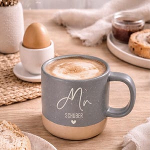 Pode incluir: Uma caneca de cer&acirc;mica cinza salpicada com uma base bege, cheia de caf&eacute; e espuma. A caneca tem uma al&ccedil;a e o nome "Mr. Sch&uuml;ber" est&aacute; escrito na frente. Um ovo cozido em uma porta-ovos e uma fatia de p&atilde;o tamb&eacute;m s&atilde;o vis&iacute;veis.
