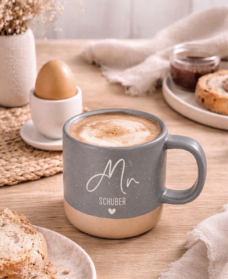 Pode incluir: Uma caneca de cer&acirc;mica cinza salpicada com uma base bege, cheia de caf&eacute; e espuma. A caneca tem uma al&ccedil;a e o nome "Mr. Sch&uuml;ber" est&aacute; escrito na frente. Um ovo cozido em uma porta-ovos e uma fatia de p&atilde;o tamb&eacute;m s&atilde;o vis&iacute;veis.
