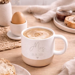 Pode incluir: Uma caneca de cer&acirc;mica branca e bege salpicada cheia de caf&eacute;, com a palavra "Mrs" e uma data impressa. A caneca faz parte de um cen&aacute;rio de caf&eacute; da manh&atilde;, com um ovo em uma x&iacute;cara, p&atilde;o e geleia.