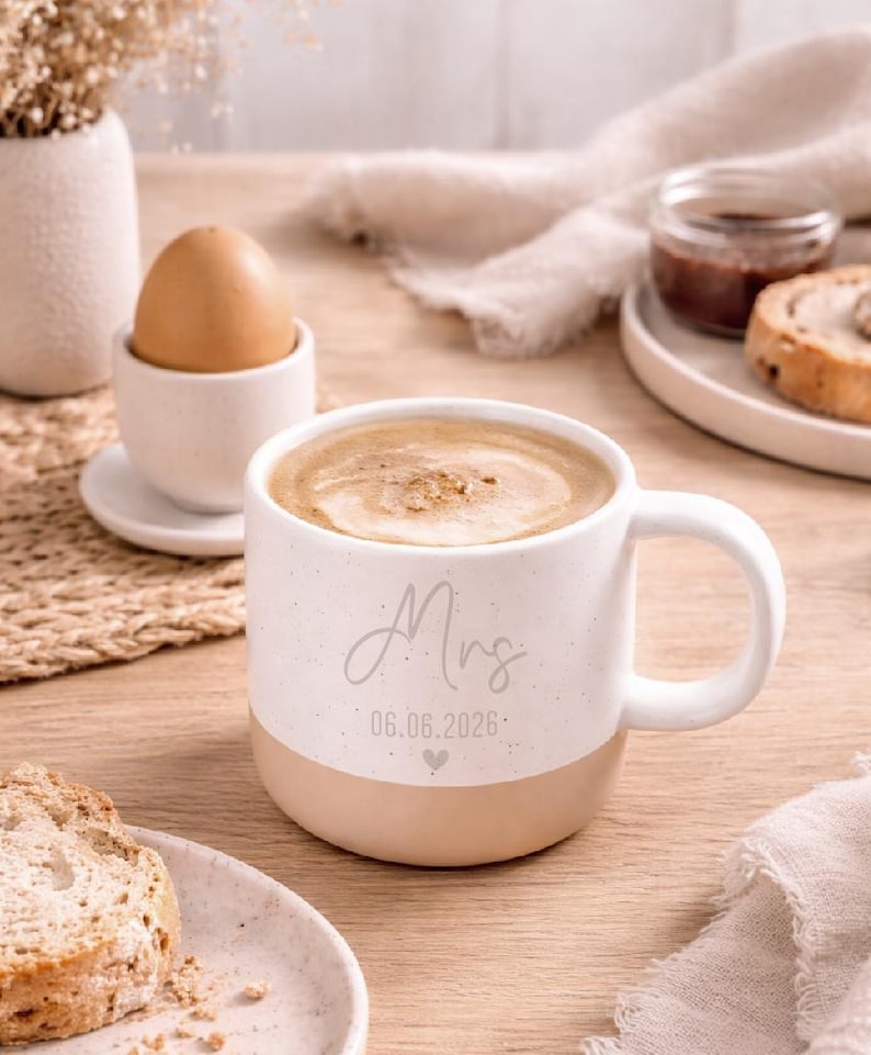 Pode incluir: Uma caneca de cer&acirc;mica branca e bege salpicada cheia de caf&eacute;, com a palavra "Mrs" e uma data impressa. A caneca faz parte de um cen&aacute;rio de caf&eacute; da manh&atilde;, com um ovo em uma x&iacute;cara, p&atilde;o e geleia.