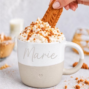 K&ouml;nnte beinhalten: Ein zweifarbiger Keramikbecher mit einem wei&szlig;en oberen Bereich, der den Namen Marie in Schreibschrift tr&auml;gt, und einem beigen unteren Bereich mit einem kleinen eingepr&auml;gten Herz. Der Becher ist mit Schlagsahne, Karamellsauce und einem Keks gef&uuml;llt.