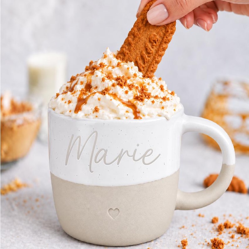 K&ouml;nnte beinhalten: Ein zweifarbiger Keramikbecher mit einem wei&szlig;en oberen Bereich, der den Namen Marie in Schreibschrift tr&auml;gt, und einem beigen unteren Bereich mit einem kleinen eingepr&auml;gten Herz. Der Becher ist mit Schlagsahne, Karamellsauce und einem Keks gef&uuml;llt.
