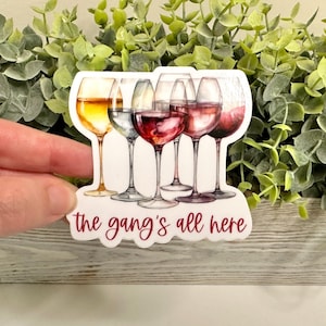 May include: White sticker with a watercolor illustration of five wine glasses, two white wine glasses and three red wine glasses. The text "the gang's all here" is written in red below the glasses.