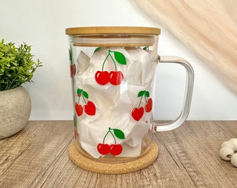 Könnte beinhalten: Eine Glas-Tasse mit einem Korkdeckel und einem Henkel. Die Tasse hat ein Kirsch-Druckdesign mit roten Kirschen und grünen Blättern.
