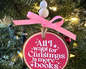 May include: A round, wooden Christmas ornament with a pink center and the text "All I want for Christmas is more books" in white. The ornament is adorned with a pink ribbon and white beads, hanging from a twine string. The ornament is hanging on a Christmas tree.