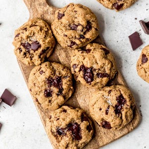 Pode incluir: Vista aérea de biscoitos com pepitas de chocolate recém-assados em uma tábua de madeira. Os biscoitos são dourados com grandes pedaços de chocolate e polvilhados com sal marinho. Pedaços de chocolate amargo espalhados.