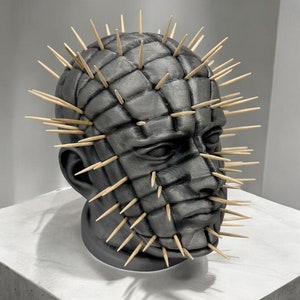 May include: A black and gray 3D printed sculpture of a human head with a grid pattern and numerous toothpicks protruding from the surface.