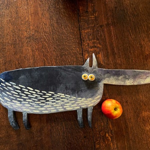 May include: A ceramic sculpture of a black dog with yellow eyes and white markings. The dog is looking to the right and has a long snout. A red apple is sitting next to the dog.