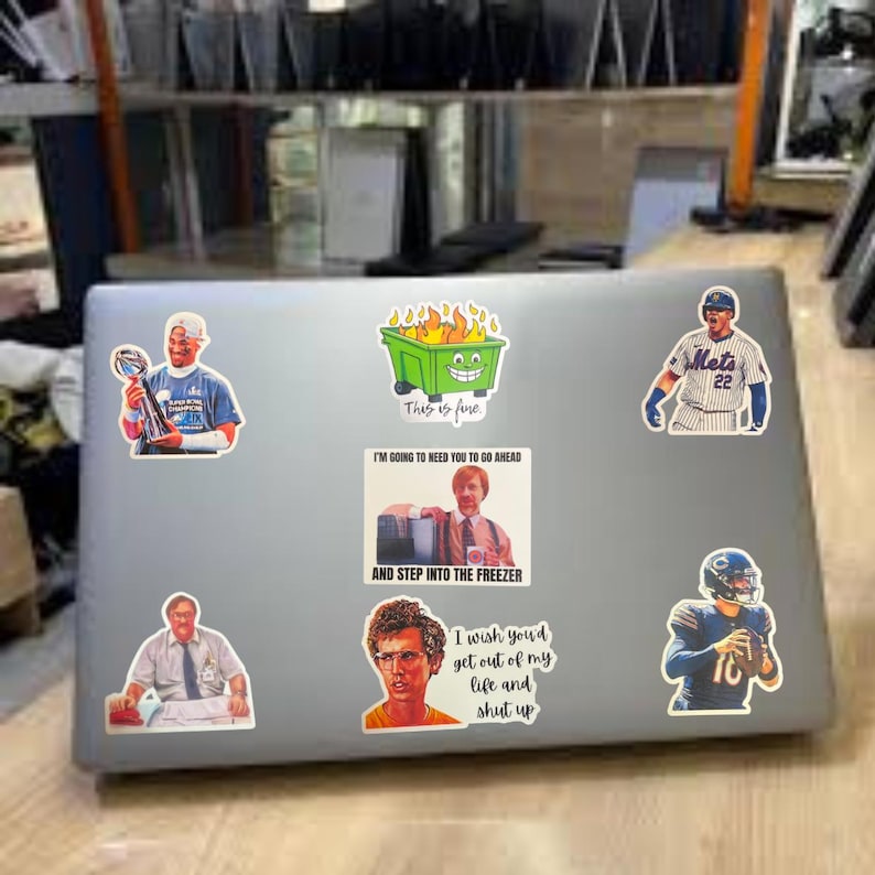 May include: A silver laptop covered in stickers. Stickers include a cartoon dumpster on fire with the text "This is fine", a baseball player, and a football player. Other stickers have text: "I'm going to need you to go ahead and step into the freezer" and "I wish you'd get out of my life and shut up."