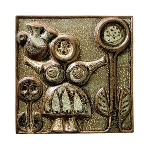 May include: Square ceramic wall art with a raised relief design. The artwork features stylized floral and organic shapes in shades of brown, green, and gold. The artist's signature "Noomi" is visible in the lower right corner.