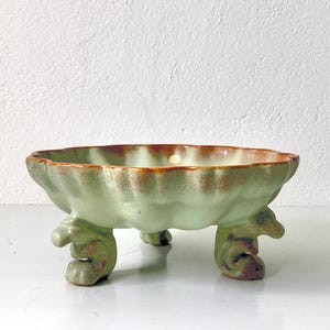 Könnte beinhalten: Eine hellgrüne Keramikschale mit gewelltem Rand und drei dekorativen Füßen. Die Schale hat einen braunen Rand und eine strukturierte Oberfläche. Die Füße haben ein einzigartiges, gewelltes Design.
