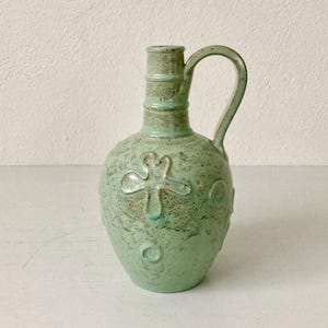 Puede incluir: Una jarra de cerámica de color verde claro con una textura marrón moteada y una sola asa. El cuerpo presenta un diseño floral en relieve de cuatro pétalos y pequeños acentos circulares. El cuello es cilíndrico con estrías horizontales.