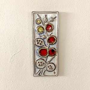 May include: A rectangular ceramic wall plaque with a white glazed surface featuring a raised botanical motif. The design includes circular elements in shades of bright red, lime green, and textured off-white, arranged along a central stem.