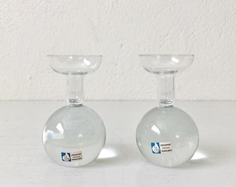 May include: Two clear glass candle holders with spherical bases and wide, shallow tops. Each piece features a small rectangular sticker on the front with the blue letter L and the text Lindshammar Sweden Gunnar Ander.
