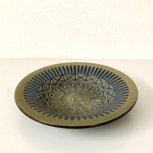 May include: An olive-green ceramic plate with a blue geometric pattern. The plate has a central circular design with radiating lines and triangles. The rim features vertical lines. The plate is on a white surface. The plate's diameter is approximately 20 cm.