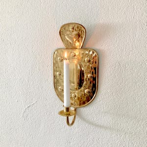 Peut inclure: Une applique murale en métal doré poli dotée d'une plaque arrière en forme de bouclier avec une texture martelée et des accents circulaires en relief. Une bougie blanche est placée dans le petit bougeoir circulaire fixé à la base.