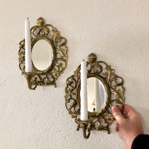 May include: Two ornate, gold-coloured wall sconces with oval mirrors and candle holders. Each sconce features a white candle. The sconces have a decorative, swirling design and are mounted on a white wall.