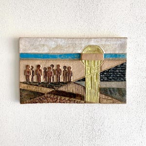 Puede incluir: Una pieza de arte mural rectangular y texturizada con un diseño en relieve. La obra presenta un paisaje estilizado con figuras, una torre y formas geométricas en tonos tierra, azul y amarillo.