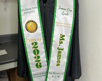 Op de afbeelding: Een witte afstudeersjerp met groene glitterrand gedragen over een zwarte afstudeertoga. De linkerkant toont het logo van Fitchburg State University, 'Class of 2026' en 'Teaching Minds Touching Hearts'. De rechterkant bevat 'Summa Cum Laude', 'Bachelors of Science Special Education', 'Ms. James' en 'In Honor of R.S.W'.