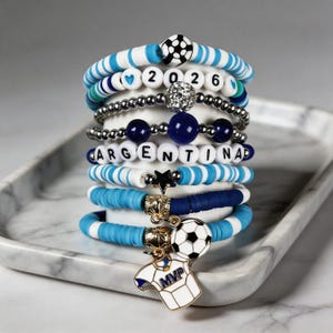 May include: Stack of beaded bracelets in shades of blue, white, and silver. Some bracelets feature the word "ARGENTINA" and the number "2026". Charms include a soccer ball and a jersey with "MVP" on it. The bracelets are stacked on a marble tray.