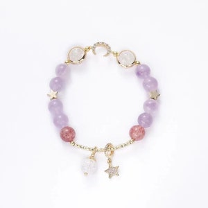 May include: A purple and pink beaded bracelet with gold accents. The bracelet features a crescent moon charm, star charms, and two round white beads with gold trim.