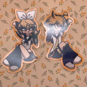 May include: Two cartoon stickers featuring anime-style characters with orange hair and outfits. One sticker depicts a female character in a sailor-style outfit, and the other shows a male character in a similar style. The stickers have a light orange border.