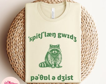 May include: A light beige t-shirt with a green raccoon illustration and the text "'spit'laen gwid3 pa'owl a djist" printed on it.