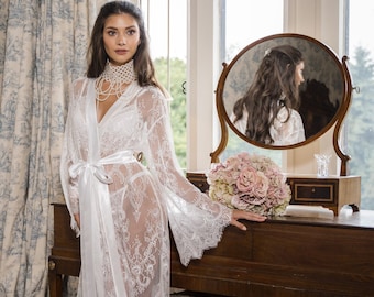 May include: A floor-length white lace robe featuring long, flared sleeves and a satin tie-waist belt. The sheer garment is worn over matching white lingerie, complemented by a white lace choker necklace and white open-toe footwear.