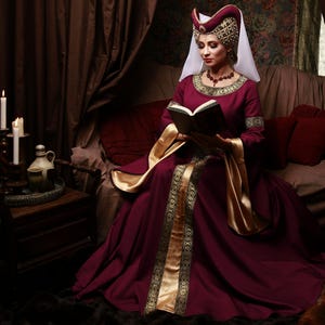 May include: A burgundy medieval-style dress with gold trim and accents. The dress features long sleeves and a flowing skirt. The person is wearing a matching burgundy and gold headdress and holding a book. The scene is set in a room with dark curtains and candles.