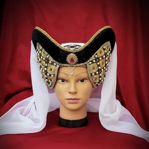 Puede incluir: Un sombrero de terciopelo negro con adornos dorados y de perlas y un velo blanco. El sombrero tiene una corona alta y puntiaguda y un ala ancha.