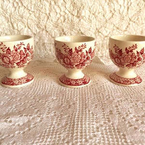 Könnte beinhalten: Drei cremefarbene Eierbecher mit rotem Blumenmuster. Die Eierbecher haben einen Sockel und eine abgerundete Schale. Das florale Design wiederholt sich auf dem Sockel und am Rand der Tasse. Sie sind auf einer weißen Spitzentischdecke angeordnet.