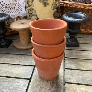 Puede incluir: Tres pequeñas macetas de color terracota apiladas sobre una superficie de madera. Las macetas tienen un diseño clásico y sencillo con un borde ligeramente ensanchado. La imagen sugiere una estética rústica y vintage, adecuada para jardinería o decoración del hogar.