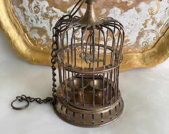 May include: A small, antique brass birdcage with a hinged door and a chain. The cage has a decorative top and a small bird figurine inside. The cage is sitting on a white surface, with a gold-framed tray in the background.