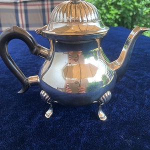 Op de afbeelding: Een zilveren theepot met een zwarte handgreep en drie poten. De theepot heeft een glanzend, reflecterend oppervlak en een koepelvormig deksel.