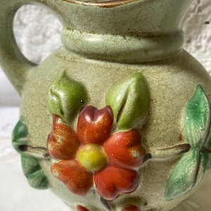 May include: A green ceramic pitcher with a floral design. The pitcher has a wide mouth and a handle. The floral design features a red flower with a yellow centre and green leaves.