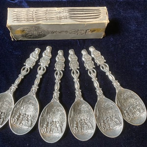 Könnte beinhalten: Sechser-Set silberne Löffel mit verzierten Griffen und detaillierten Gravuren einer historischen Szene. Die Löffel befinden sich in ihrer Originalverpackung.