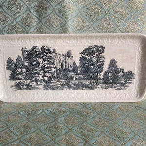 Puede incluir: Una bandeja de servir de cerámica blanca con una imagen en blanco y negro de un castillo y árboles. La bandeja tiene un borde elevado y un borde decorativo.