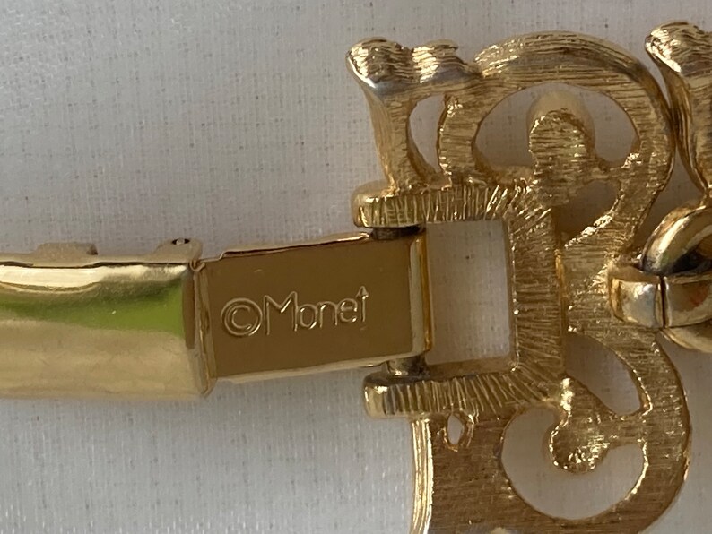 May include: A gold-toned metal bracelet with a clasp that reads "&copy;Monet". The bracelet has a decorative, ornate design.