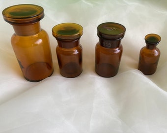 May include: Four amber glass bottles with cork stoppers. The largest bottle is taller and wider than the other three. The three smaller bottles are all the same size. The bottles are all empty and have a vintage look.