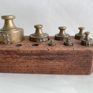 Könnte beinhalten: Ein Satz Messinggewichte in einer Holzkiste. Die Gewichte sind unterschiedlich groß und haben eine kreisförmige Form. Die Kiste ist braun und hat eine Holzmaserung.