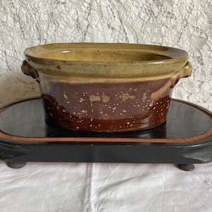 Puede incluir: Un macetero de cerámica esmaltada marrón y amarillo con dos asas. El macetero está sobre un soporte de madera negro.