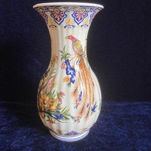 May include: A white ceramic vase with a blue and yellow floral design and a bird with a long tail. The vase has a fluted body and a wide mouth.