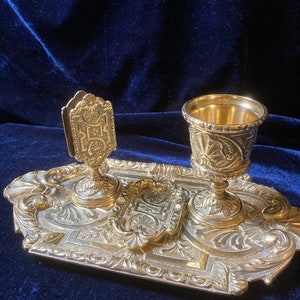 Pode incluir: Um conjunto de secretária dourado com um design ornamentado. O conjunto inclui um porta-canetas, um tinteiro e uma bandeja. A bandeja tem uma borda elevada e é decorada com um padrão floral.